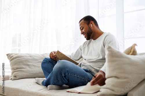 Smiling african american man in earphone reading book near notebook on couch at home, self-improvement concept
