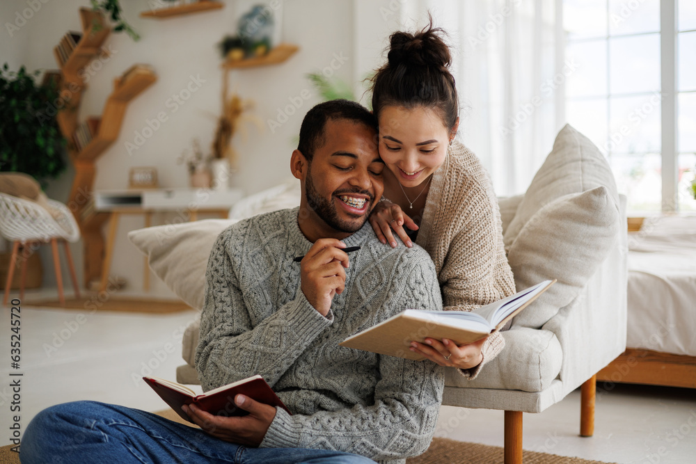 Joyful multiracial couple in warm clothes reading book together while spending time at home - obrazy, fototapety, plakaty 