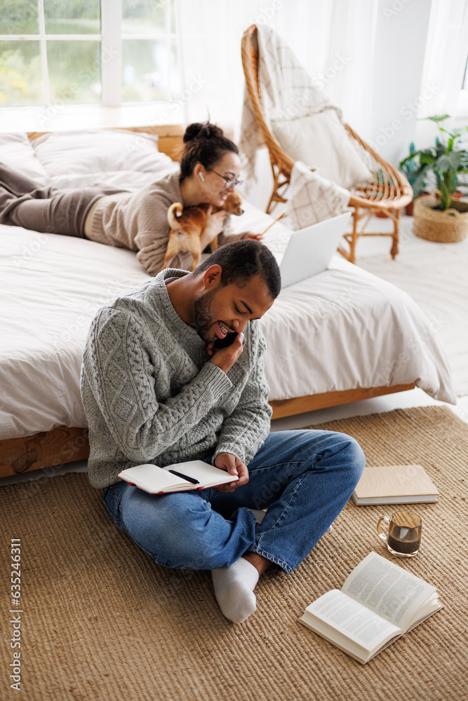 Obraz premium Smiling african american man talking on smartphone near books, coffee and girlfriend with dog at home