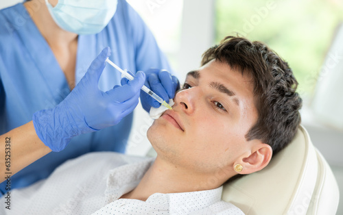 Young male patient lying on clinical chair undergoing face care procedure by injection method