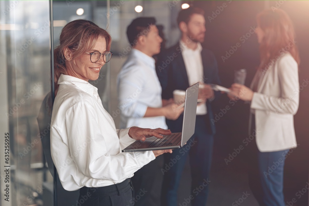 Obraz premium Mature smiling businesswoman at office with group of colleagues on background, working on laptop