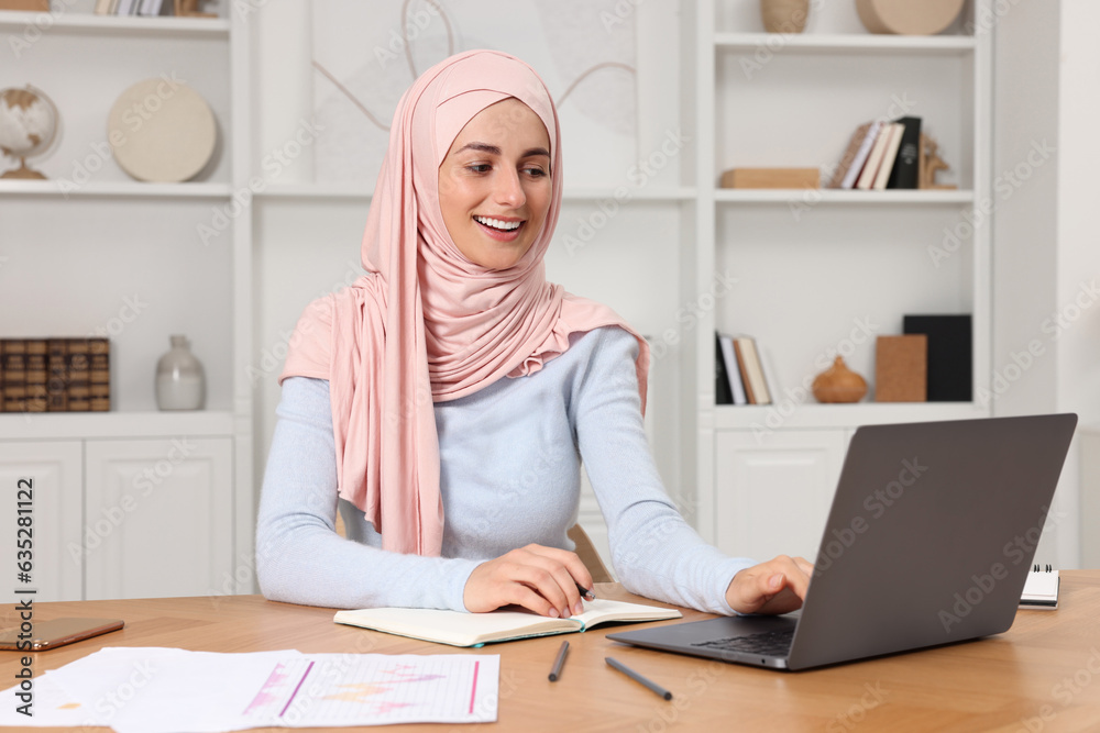 Muslim woman writing notes near laptop at wooden table in room Muslim woman writing notes near laptop at wooden table in room