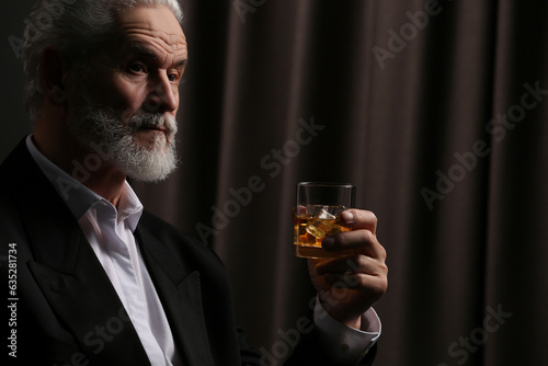 Senior man in suit holding glass of whiskey with ice cubes on brown background. Space for text