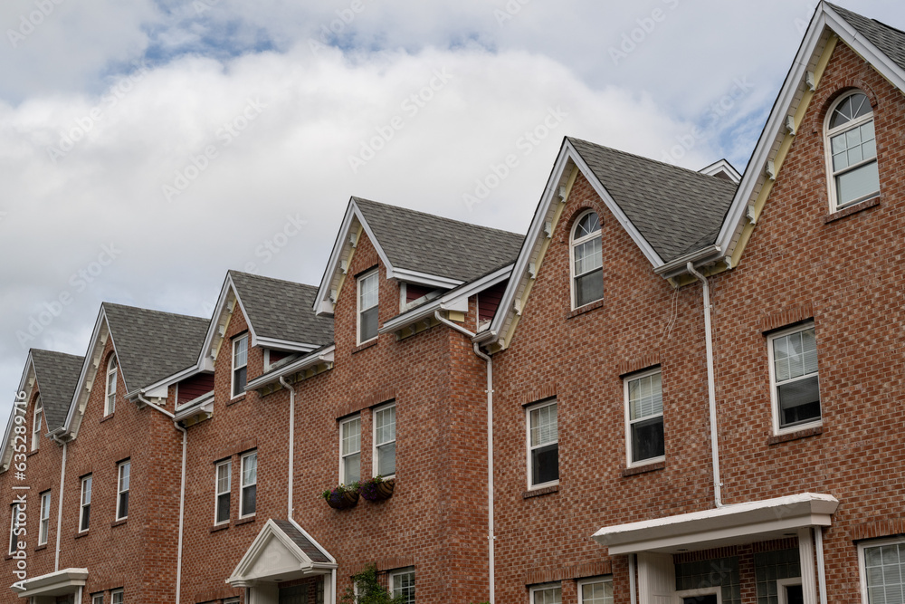 A row of brown brick townhouses with peaked gable roofs, and double hung windows with white trim. The shingles are black asphalt. The adjoined facade row houses are multi-story residential buildings. A row of brown brick townhouses with peaked gable roofs, and double hung windows with white trim. The shingles are black asphalt. The adjoined facade row houses are multi-story residential buildings.