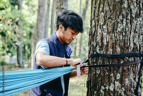 Young Asian man tying rope of hammock to tree in forest. Camping and travel concept.
