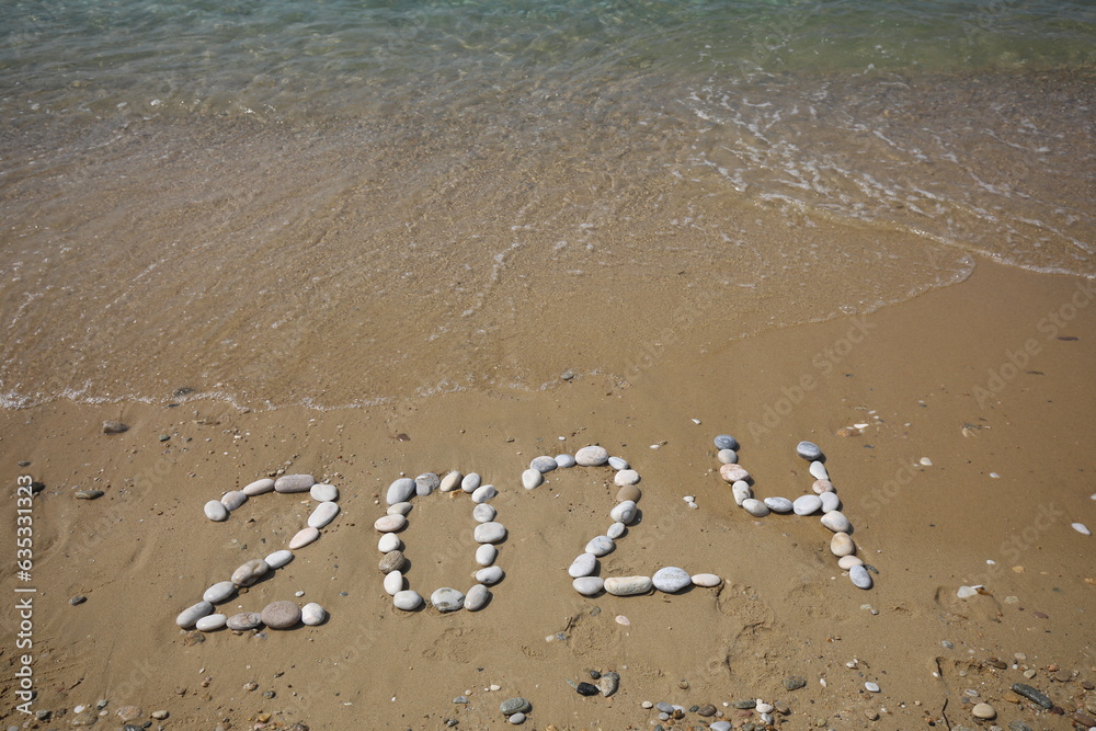Fototapeta premium New Year 2024, lettering on the beach with wave and clear blue sea. Numbers 2024 year on the sea shore, message handwritten in the golden sand on beautiful beach background. New Years concept.