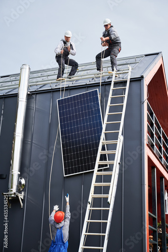Men workers installing solar panel system on roof of house. Electricians in helmets lifting up photovoltaic solar module with help of ropes outdoors. Concept of alternative and renewable energy.