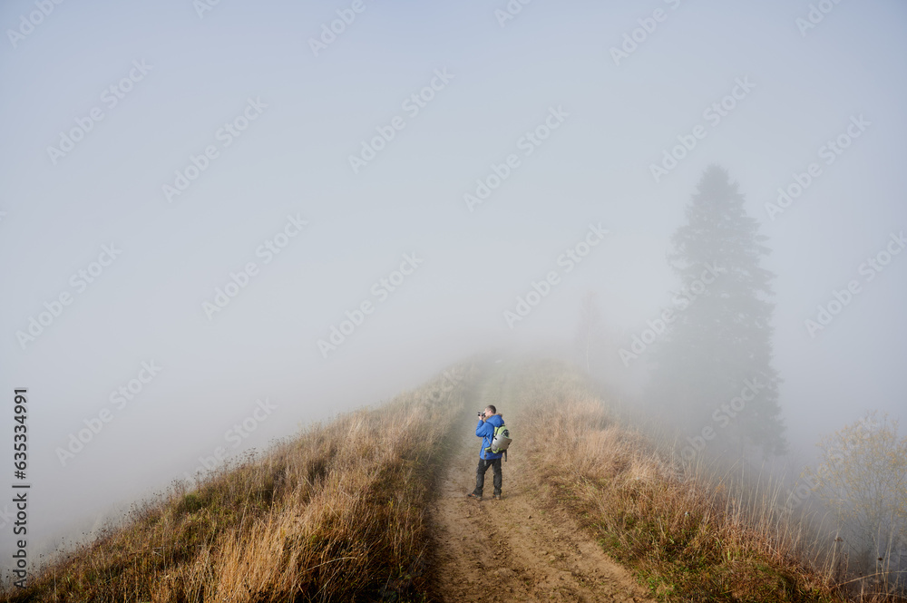 Photographer taking picture with professional camera while standing on grassy hill road with dense fog on background