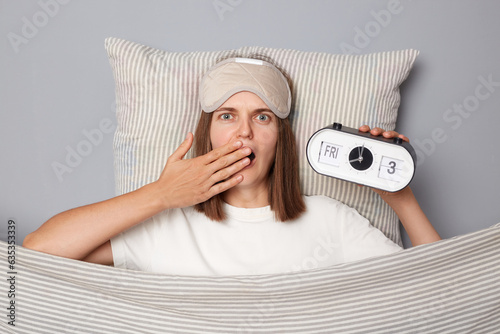 Shocked woman in white T-shirt and sleeping eye mask lie in bed on pillow under blanket isolated on gray background holding alarm looking with scared expression photo