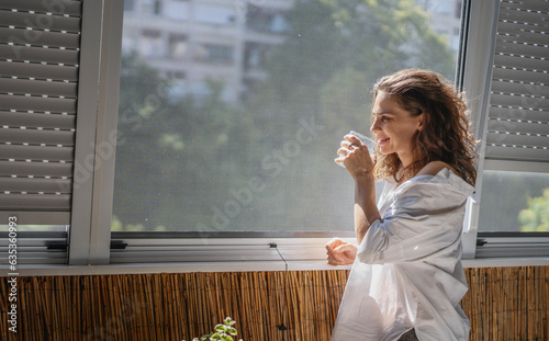 Obraz na płótnie Young smiling caucasian woman in a white shirt standing on the balcony in the morning drinking water and meeting a new day w salonie