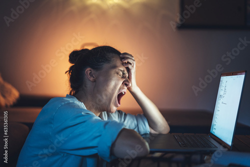 Young woman yawning in front of laptop screen while working from home at night. Deadline and overworking photo