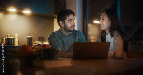 Portrait of Young Couple Using Laptop Computer While Sitting in the Kitchen at Home And Talking. Engaged Couple Doing Online Shopping for their Wedding Preparations, Discussing Different Options.