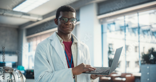 Portrait of Black Young Male Scientist Using Laptop Computer, Looking at Camera and Smiling in a Laboratory. Professional African American Man Working as Engineer, Monitoring Technology Projects
