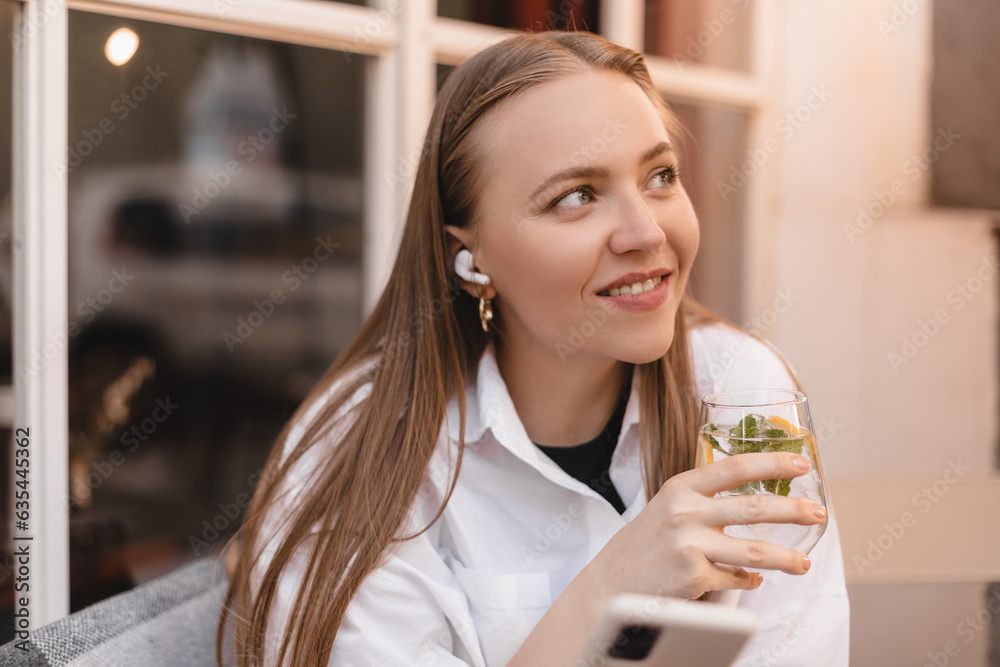 Beautiful happy girl in wireless earphones drinking water with lemon and mint while using mobile phone outdoor cafe. Beautiful happy girl in wireless earphones drinking water with lemon and mint while using mobile phone outdoor cafe.