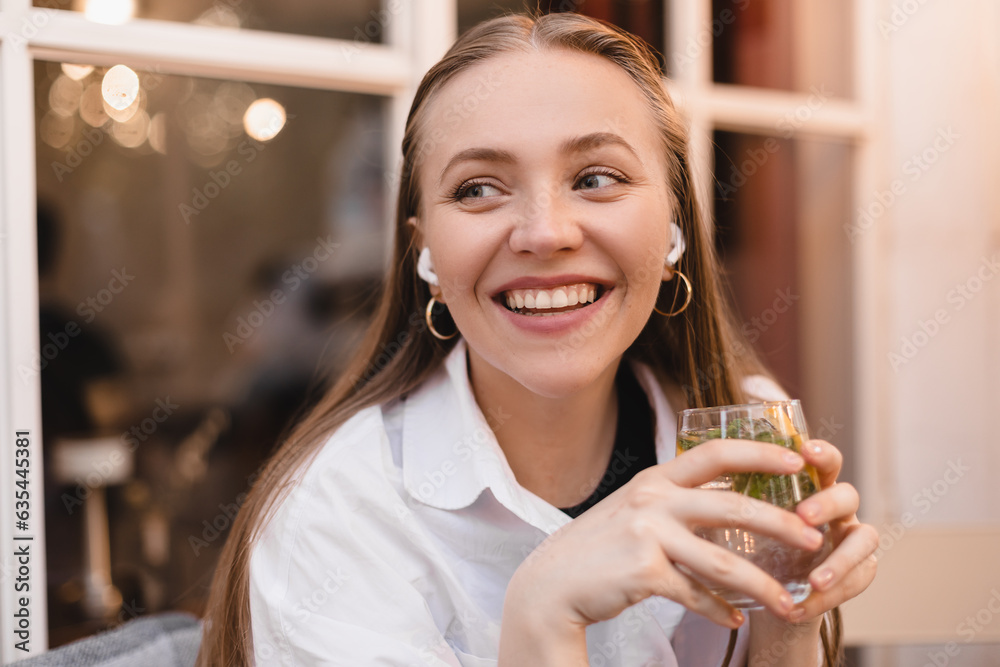 Close up smiling young woman is sitting in a restaurant and hold glass water, lemon, mint. Woman is relaxing in wireless headphones. A young woman in restaurant using technologies listening music.