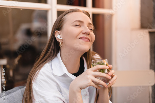 Closed eyes smiling young woman is sitting in a restaurant and hold glass water, lemon, mint. Woman is relaxing in wireless headphones. Young woman in restaurant using technologies listening music.