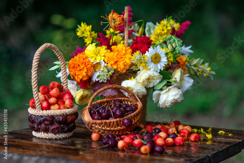 Artistic still life with a bouquet of garden flowers and cherries in a basket on a wooden table against a dark background