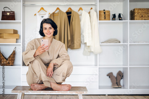 Woman sit in front of open wardrobe at home, choosing trendy clothes via smartphone photo