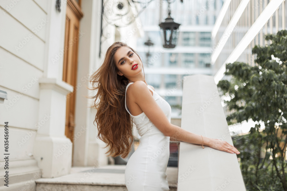 Fototapeta premium Glamorous beautiful woman model with brunet hairstyle in fashionable elegant white dress stands and poses near a white vintage building on the street
