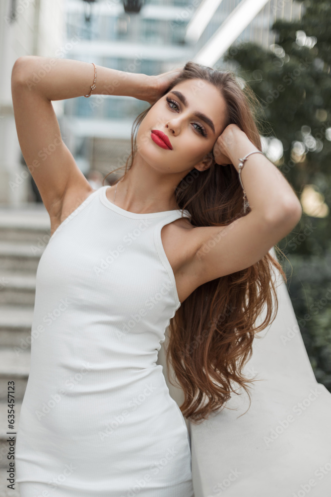 Tender beautiful sensual girl model with red lips in a fashionable elegant dress straightens her hair and walks in the city
