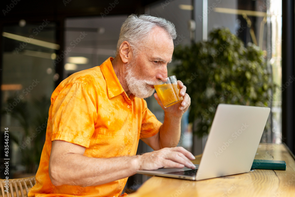 Senior man using laptop, working at workplace, drinking cocktail or lemonade, remote work in office Senior man using laptop, working at workplace, drinking cocktail or lemonade, remote work in office