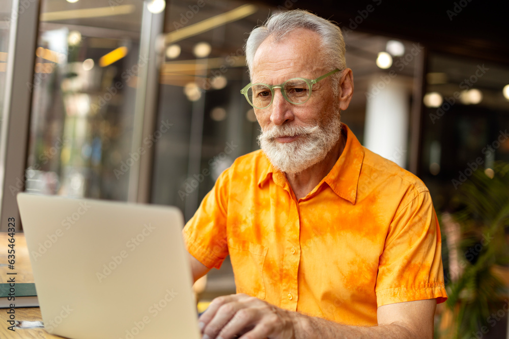 Smiling mature freelancer wearing glasses wearing stylish orange shirt, using laptop, working online Smiling mature freelancer wearing glasses wearing stylish orange shirt, using laptop, working online