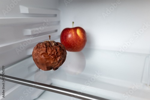 Moldy apple on the shelf of an open refrigerator. Two apples, moldy in the foreground and ripe in the background. Expired food, spoiled food.