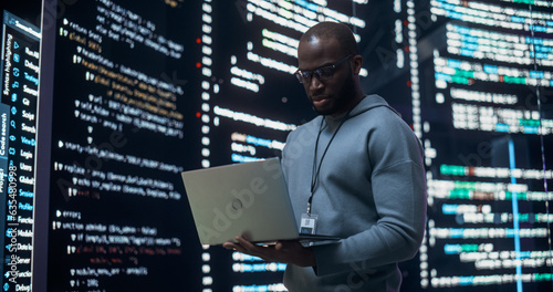 Portrait of Young Black Man Working on Laptop, Looking at Big Digital Screen Displaying Back-end Code Lines. Professional Programmer Developing a Big Data Interface Software Project Concept.