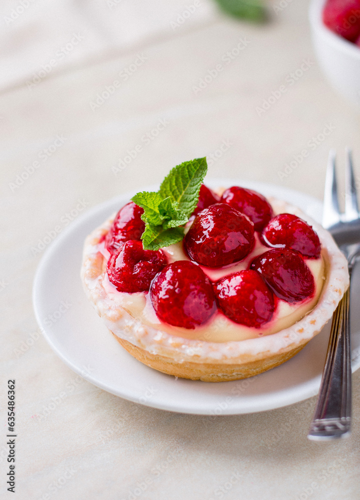 A plate with a custard cake, decorated with raspberries and a mint leaf, on a light background, close up. A plate with a custard cake, decorated with raspberries and a mint leaf, on a light background, close up.