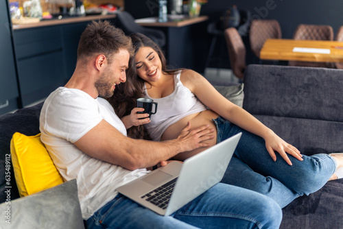 Expecting new parents are sitting on the couch planning the decor for the new children's room, the laptop is on his lap while he is touching her belly,she holds a cup of tea in her hand