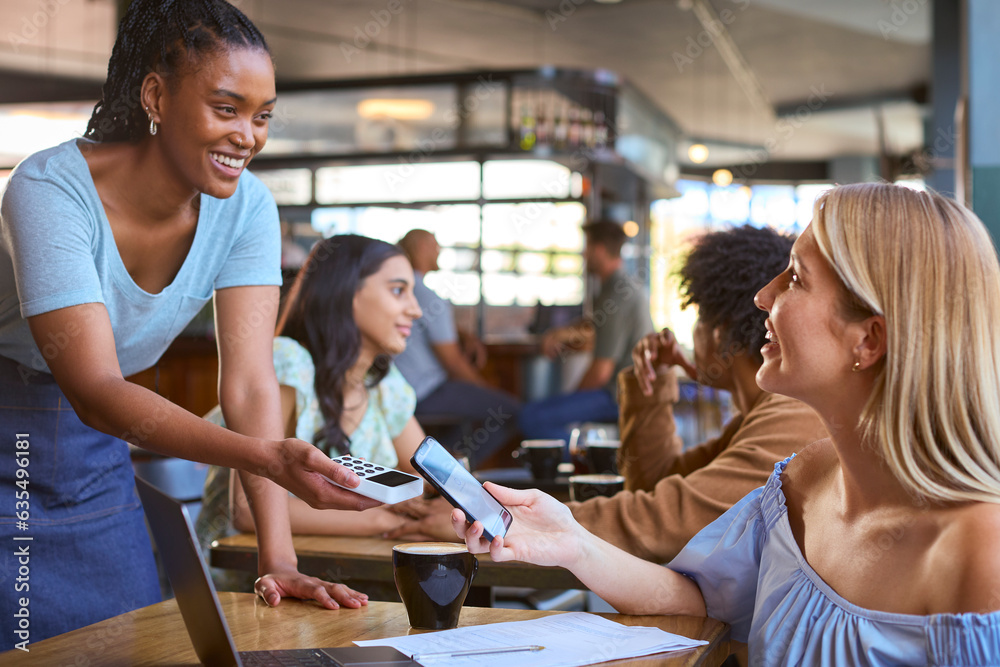 © Monkey Business - Woman In Coffee Shop Working On Laptop Paying Bill With Contactless Mobile Phone Payment