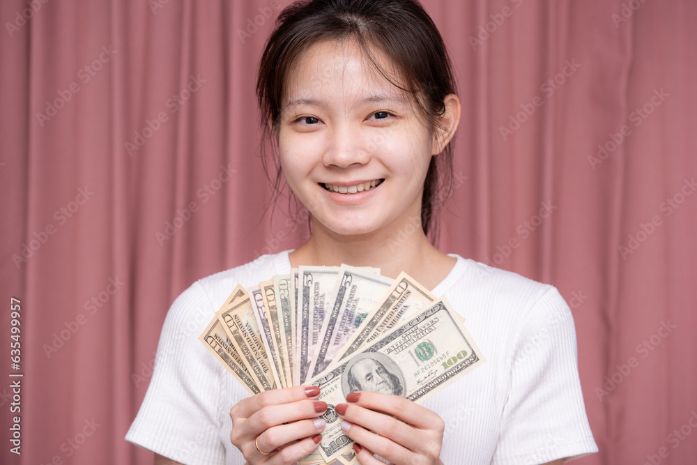 Portrait of a smiling Asian woman wear white shirt and holding banknotes or money us dollar on pink background Portrait of a smiling Asian woman wear white shirt and holding banknotes or money us dollar on pink background