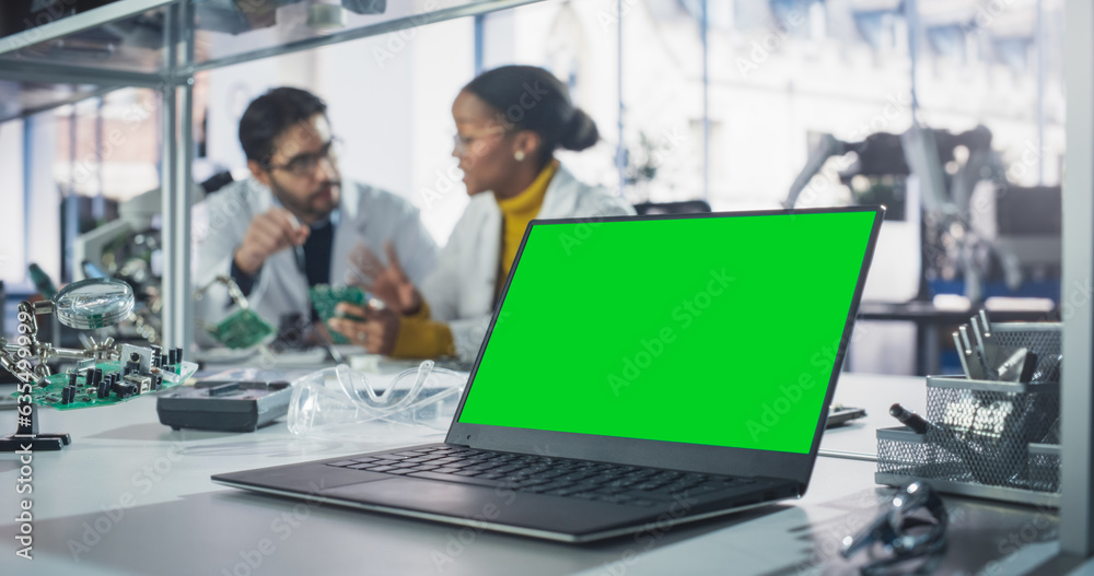 Laptop Computer with Green Screen Mock Up Display Standing on a Table in a Science Class Laboratory. Diverse University Graduates Having a Conversation About Their Electronics Project Laptop Computer with Green Screen Mock Up Display Standing on a Table in a Science Class Laboratory. Diverse University Graduates Having a Conversation About Their Electronics Project