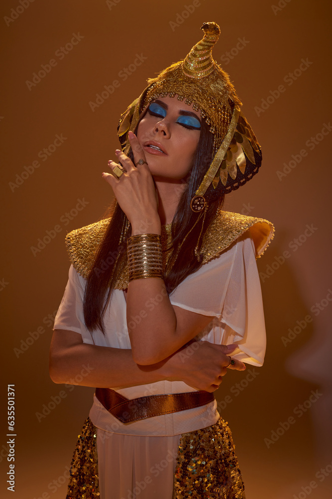 Stylish woman with bold makeup and Egyptian attire posing with closed eyes on brown background