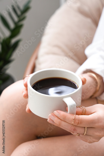 Young woman sitting in armchair and holding cup of delicious coffee