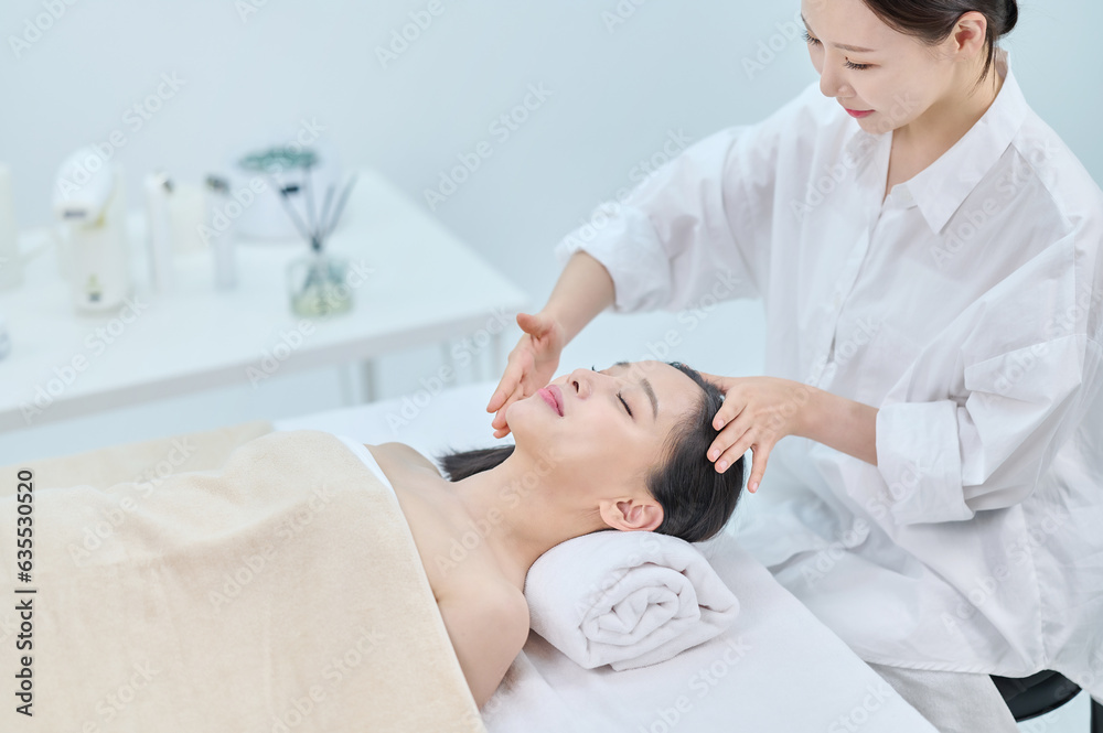 Obraz premium 아시아 한국의 젊은 여성이 스파 또는 피부관리전문점 또는 피부과 병원에서 침대에 누워 베이지색 대형 타올을 덮고 눈을 감고있고 하얀색 셔츠를 입은 전문관리사가 손으로 얼굴 피부관리 또는 두피관리를 받고 있다. 