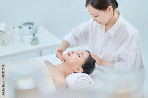 아시아 한국의 젊은 여성이 스파 또는 피부관리전문점 또는 피부과 병원에서 침대에 누워 베이지색 대형 타올을 덮고 눈을 감고있고 하얀색 셔츠를 입은 전문관리사가 손으로 얼굴 피부관리 또는 두피관리를 받고 있다.  photo