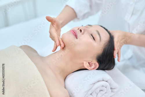 아시아 한국의 젊은 여성이 스파 또는 피부관리전문점 또는 피부과 병원에서 침대에 누워 베이지색 대형 타올을 덮고 눈을 감고있고 하얀색 셔츠를 입은 전문관리사가 손으로 얼굴 피부관리 또는 두피관리를 받고 있다.  photo
