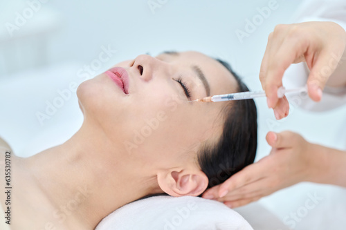 아시아 한국의 젊은 여성이 피부과 병원 또는 성형외과의 침대에 누워 베이지색 대형 타올을 덮고 눈을 감고 있고 하얀색 셔츠를 입은 전문의사가 주사기를 들고 충전제 또는보톡스로 노화방지 또는 얼굴 주름을 관리하고 있다. 