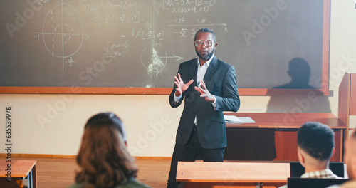 African American male professor handing out papers with tasks for test exam to muliethnic students. Examination at University concept. Man teacher giving exercises or materials for studying at lesson. photo