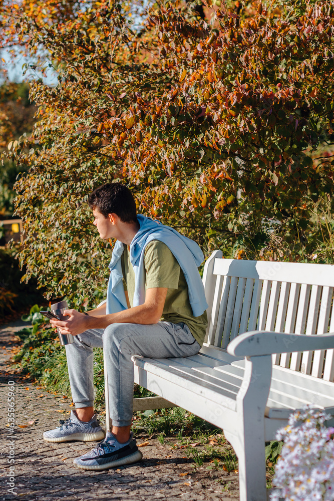 A Teenager Sits On A Bench In The Autumn Park Drinks Coffee From A Thermo Mug And Looks Into A Phone. Portrait Of Handsome Cheerful Guy Sitting On Bench Fresh Air Using Device Browsing Media Smm A Teenager Sits On A Bench In The Autumn Park Drinks Coffee From A Thermo Mug And Looks Into A Phone. Portrait Of Handsome Cheerful Guy Sitting On Bench Fresh Air Using Device Browsing Media Smm