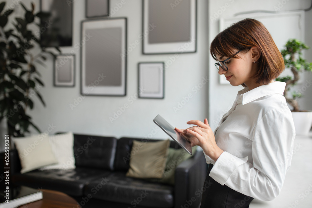 Pretty european businesswoman chatting on digital tablet in office interior, standing indoors, free space Pretty european businesswoman chatting on digital tablet in office interior, standing indoors, free space