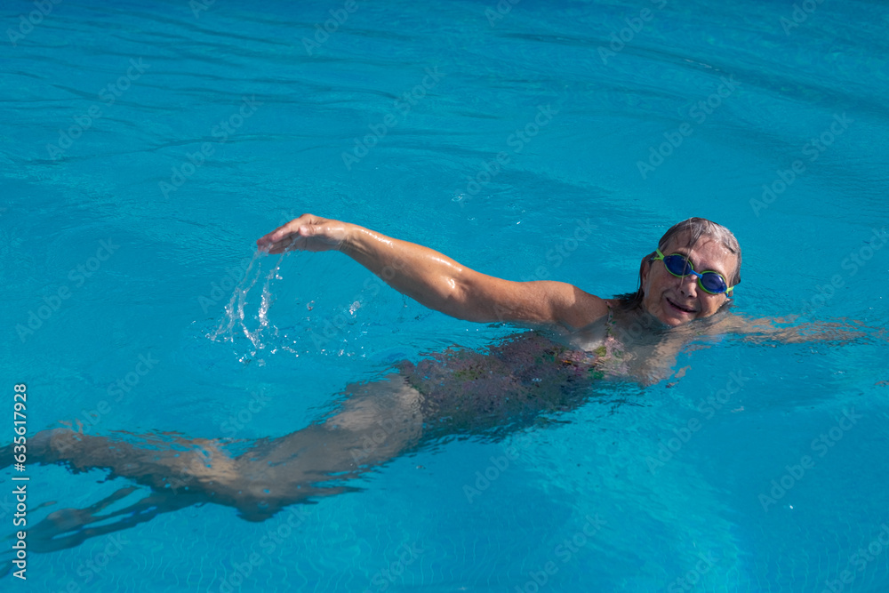 Active senior caucasian woman swimming freestyle in outdoor swimming pool under the sunlight wearing swimming goggles. Retirement and healthy lifestyle concept