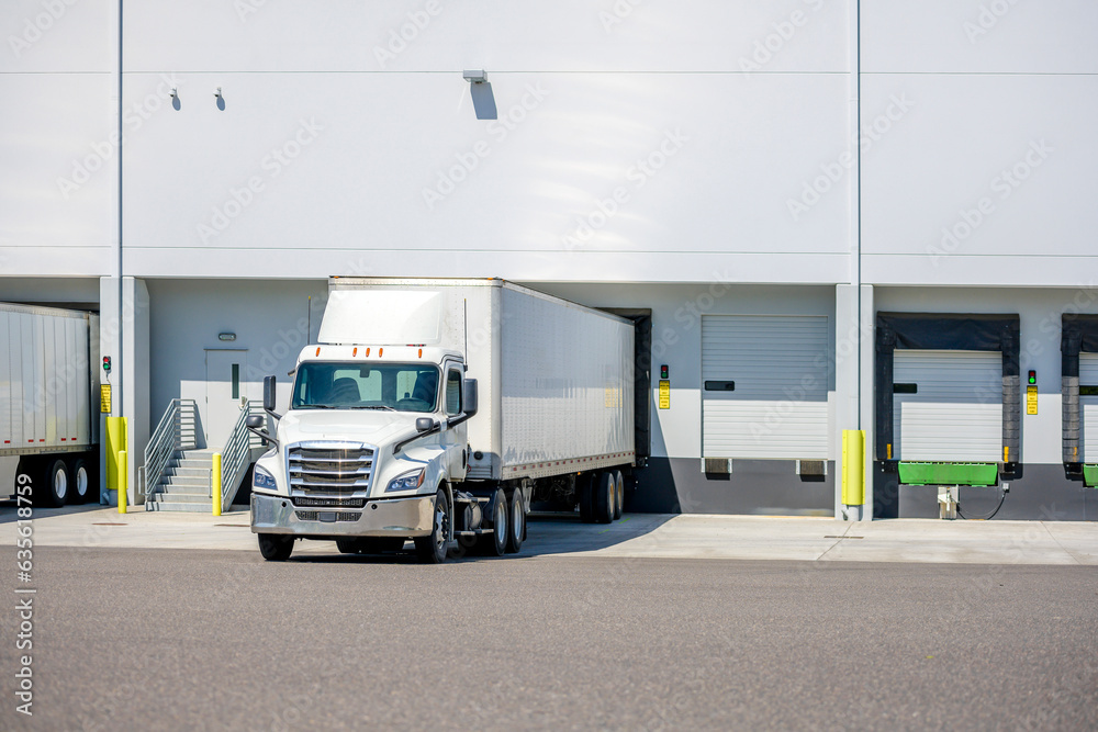 © vit - Day cab white big rig semi truck with dry van semi trailer standing on the warehouse dock gate loading cargo for the next commercial delivery