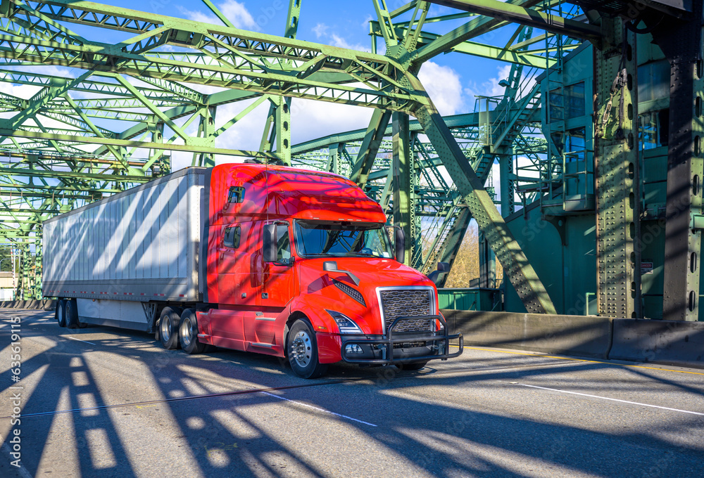 Fototapeta premium Long haul bright red big rig semi truck transporting cargo in dry van semi trailer running on the arched truss bridge