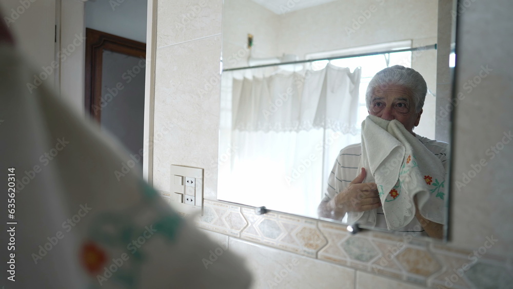 Senior man drying face with bathroom towel standing in front of mirror looking at his own reflection. Older person contemplating old age while in morning routine ritual