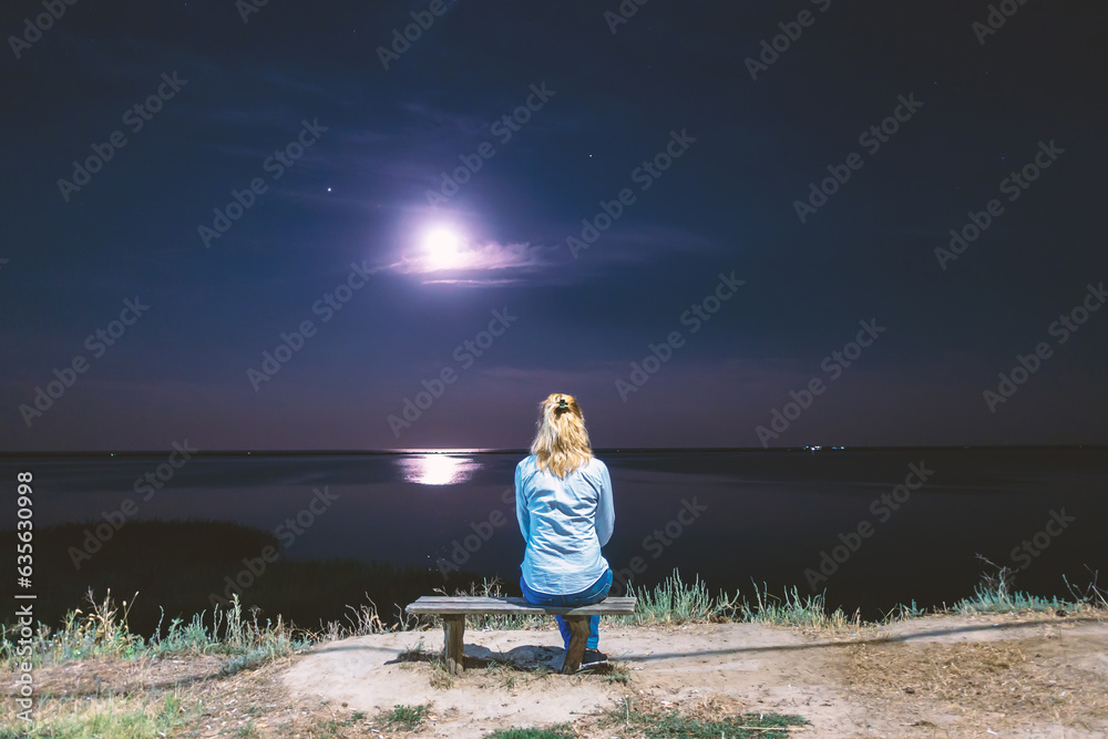 A girl sits by the sea at night under the moon