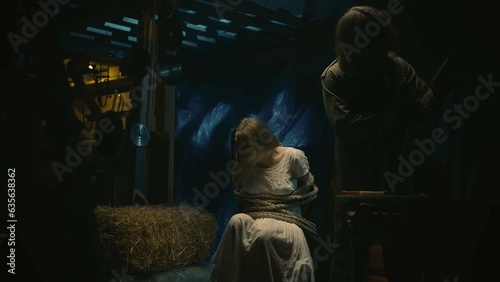 Foto Female model tied with a rope sitting on a chair, maniac picking the tools for murder