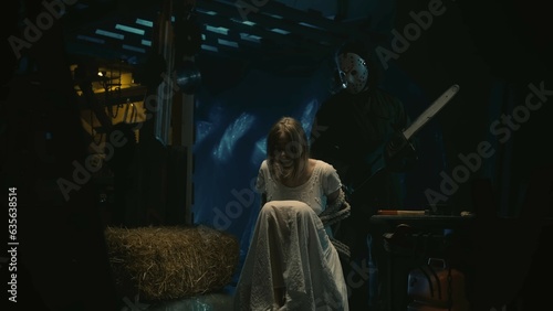 Female tied with a rope on a chair in warehouse, man in the mask with a chainsaw standing behind her.