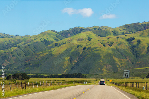 Road stretches into the distance against the backdrop of green meadows and mountains. Asphalt road with a dividing strip goes into the distance. Car drive far along the highway, rear view